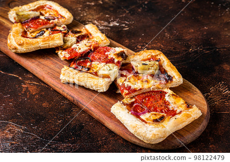 Baked Puff pastry tart pizza with artichoke, mozzarella, tomatoes and cheese sliced on a wooden board. Dark background. Top view 98122479