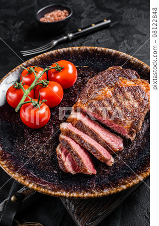 Grilled rib eye steak, ribeye beef marbled meat on a plate with tomato. Black background. top view Grilled rib eye steak, ribeye beef marbled meat on a plate with tomato. Black background. top view 98122848