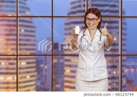 Young female doctor in glasses is showing medicine bottle and giving thumb up. Checkered windows with night cityscape view. Young female doctor in glasses is showing medicine bottle and giving thumb up. Checkered windows with night cityscape view. 98123744
