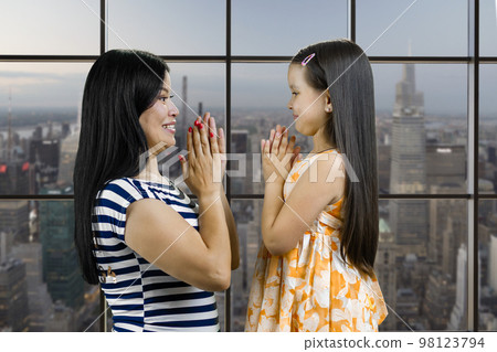 Side view of a young mother and her daughter are playing clapping game. Checkered window background with cityscape view. Side view of a young mother and her daughter are playing clapping game. Checkered window background with cityscape view. 98123794