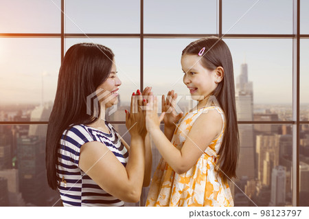 Side view of woman and her daughter are playing clapping game. Checkered window background with cityscape view. Side view of woman and her daughter are playing clapping game. Checkered window background with cityscape view. 98123797