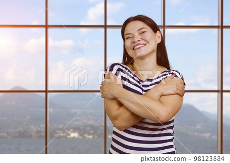 Portrait of happy young asian woman hugging herself. Checkered windows with view on mountain landscape on background. Portrait of happy young asian woman hugging herself. Checkered windows with view on mountain landscape on background. 98123884