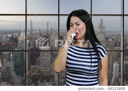 Portrait of a young asian woman is sneezing in handkerchief. Checkered windows background with cityscape view. 98123889