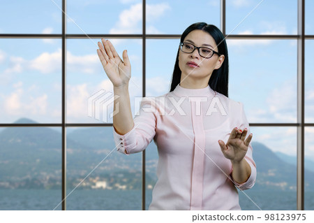 Young asian businesswoman is swiping something on imaginary virtual screen by her hand. Checkered windows with mountainscape background. 98123975