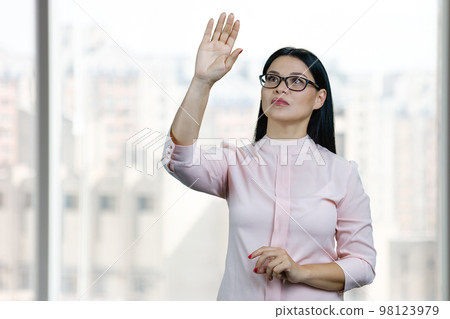 Young asian business woman is touching invisible virtual screen by her hand. Blurred windows background. 98123979