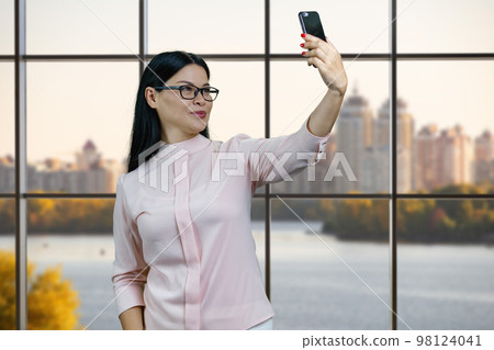 Young asian woman is taking a selife on her smartphone. Window with evening cityscape on background. 98124041