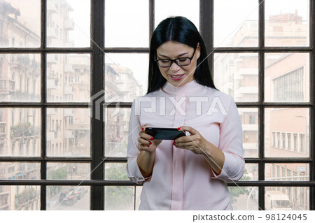 Young asian businesswoman watching a video on her smartphone device. Checkered windows background. Young asian businesswoman watching a video on her smartphone device. Checkered windows background. 98124045