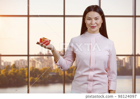 Portrait of young asian businesswoman holding gift box. Checkered windows with cityscape on background. Portrait of young asian businesswoman holding gift box. Checkered windows with cityscape on background. 98124097