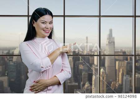 Portrait of young asian woman pointing at windows with cityscape. Happy chineese businesswoman showing copy space. Portrait of young asian woman pointing at windows with cityscape. Happy chineese businesswoman showing copy space. 98124098
