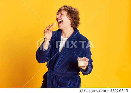 Laughing young man wearing bathrobe and having breakfast isolated over bright yellow background. Positive emotions, home cozy atmosphere, funny meme emotions 98126545