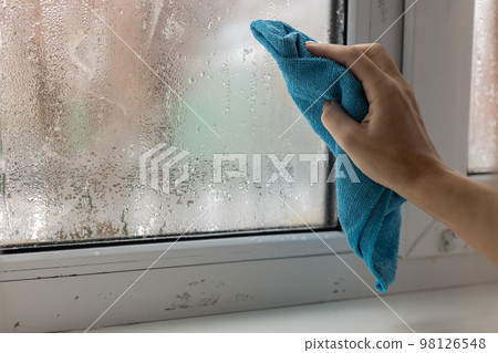 A womans hand wipes the condensate from the window glass with a towel. A womans hand wipes the condensate from the window glass with a towel. 98126548
