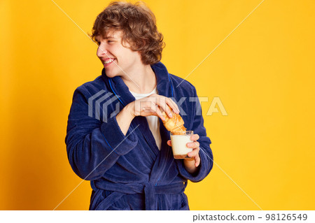Morning. Emotional young man wearing bathrobe and having breakfast isolated over bright yellow background. Positive emotions, home cozy atmosphere, funny meme emotions Morning. Emotional young man wearing bathrobe and having breakfast isolated over bright yellow background. Positive emotions, home cozy atmosphere, funny meme emotions 98126549