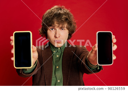 Studio footage young man, student or office clerk in vintage fashion style costume showing phone screen at camera isolated on dark red background. 98126559