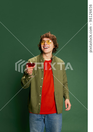 Portrait of Caucasian young handsome man in casual style clothes standing with wine glass against green background. Love, St. Valentine's day, greetings concept 98126656
