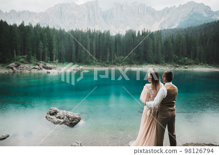Wedding couple on the nature is hugging each other near a beautiful lake in the mountains.. Beautiful model girl in white dress. Man in suit Wedding couple on the nature is hugging each other near a beautiful lake in the mountains.. Beautiful model girl in white dress. Man in suit 98126794