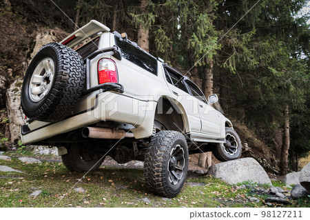 White off-road car in travel on mountain forest, Tuned reinforced suspension helps to overcome obstacles White off-road car in travel on mountain forest, Tuned reinforced suspension helps to overcome obstacles 98127211