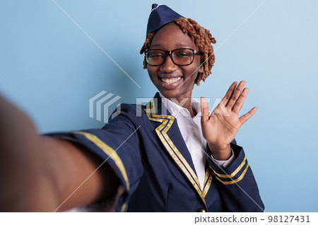 African american flight attendant waving happily in studio selfie. Aviation worker wearing glasses, afro woman taking self portrait with cellphone, smartphone. Stewardess portrait greeting. 98127431