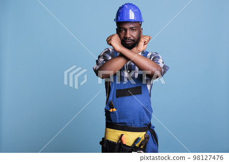 Disapproving african american builder making stop gesture with crossed arms while standing on blue background. Construction worker showing deny hands symbol while refusing to accept more work. 98127476