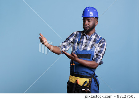 Construction worker wearing hard hat pretending to hold advertising banner in studio shot against blue background. Builder with arms raised as if carrying something and looking intently. Construction worker wearing hard hat pretending to hold advertising banner in studio shot against blue background. Builder with arms raised as if carrying something and looking intently. 98127503