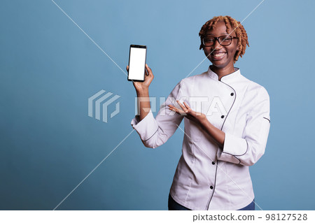 Young curly haired, kitchen chef with glasses, happily showing recipes on smartphone screen. Young African american woman, professional cooker holding portable cell phone. Young curly haired, kitchen chef with glasses, happily showing recipes on smartphone screen. Young African american woman, professional cooker holding portable cell phone. 98127528