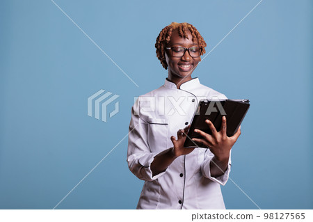 Female chef with digital tablet standing in studio while looking up recipes for new menu items. Head chef with mobile device providing garnish ideas for a gourmet cuisine meal. Female chef with digital tablet standing in studio while looking up recipes for new menu items. Head chef with mobile device providing garnish ideas for a gourmet cuisine meal. 98127565