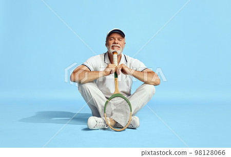 Portrait of handsome senior man in stylish white outfit sitting, leaning head on tennis racket over blue background. Concept of leisure activity, hobby, lifestyle 98128066