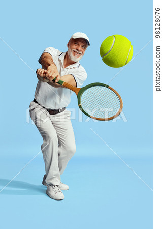 Returning ball. Portrait of handsome senior man in stylish white outfit playing tennis over blue background. Concept of leisure activity, hobby, lifestyle 98128076