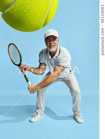 Serving ball. Portrait of handsome senior man in stylish white outfit playing tennis over blue background. Concept of leisure activity, hobby, lifestyle Serving ball. Portrait of handsome senior man in stylish white outfit playing tennis over blue background. Concept of leisure activity, hobby, lifestyle 98128081