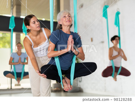 Supportive female trainer helping to mature female during antigravity aerial yoga exercise in studio 98128288