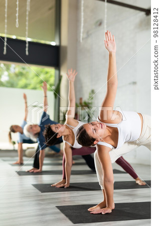 Calm young woman practicing Extended Side Angle Pose Utthita Parsvakonasana during group yoga class in studio Calm young woman practicing Extended Side Angle Pose Utthita Parsvakonasana during group yoga class in studio 98128412