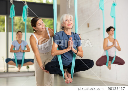 Supportive female trainer helping to mature female during antigravity aerial yoga exercise in studio Supportive female trainer helping to mature female during antigravity aerial yoga exercise in studio 98128437