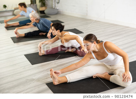 Concentrated young girl making stretching legs with group of active adult women sitting on yoga mat and doing exercise yoga Concentrated young girl making stretching legs with group of active adult women sitting on yoga mat and doing exercise yoga 98128474