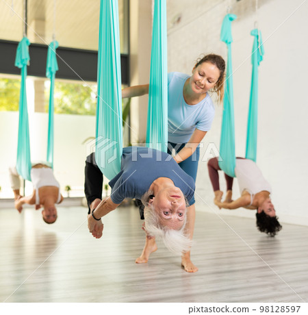 Supportive female trainer helping to mature female during antigravity aerial yoga exercise in studio Supportive female trainer helping to mature female during antigravity aerial yoga exercise in studio 98128597