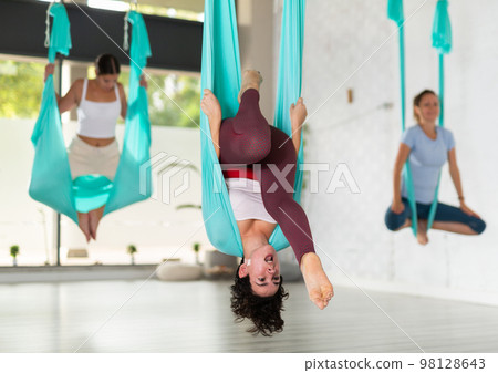 Beautiful chilean young woman hanging upside down while practicing aerial yoga during group class in fitness club Beautiful chilean young woman hanging upside down while practicing aerial yoga during group class in fitness club 98128643