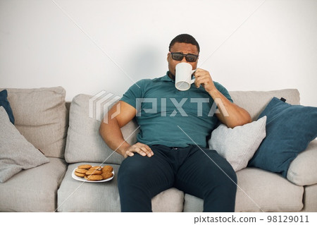 Black man sitting on a coach and holding a cup with a tea. Man wearing blue t-shirt and 3D glasses. Man watching a movie. 98129011