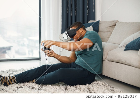 Black man in vr glasses, playing video games with virtual reality headset. Man wearing blue t-shirt. Man in virtual reality glasses sitting at home near coach. Black man in vr glasses, playing video games with virtual reality headset. Man wearing blue t-shirt. Man in virtual reality glasses sitting at home near coach. 98129017