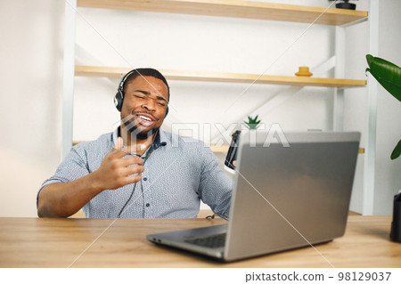 Black business man sitting in office with a laptop. Bearded man in earphones has a video call. Man wearing blue shirt and holding takeaway coffee. Black business man sitting in office with a laptop. Bearded man in earphones has a video call. Man wearing blue shirt and holding takeaway coffee. 98129037