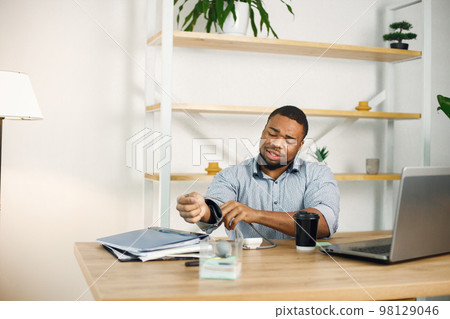 Black business man sitting in office with a laptop. Bearded man measuring arterial pressure with tonometr. Man wearing blue shirt. 98129046