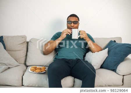Black man sitting on a coach and holding a cup with a tea. Man wearing blue t-shirt and 3D glasses. Man watching a movie. Black man sitting on a coach and holding a cup with a tea. Man wearing blue t-shirt and 3D glasses. Man watching a movie. 98129064
