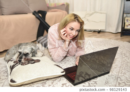 Lovely woman in a pink sweater is lying on the carpet with a laptop at home, a gray dog is sleeping on a pillow next to her. Lovely woman in a pink sweater is lying on the carpet with a laptop at home, a gray dog is sleeping on a pillow next to her. 98129143