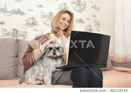 Gray shaggy dog is sitting on the couch in the arms of a girl who is working on a laptop. 98129144