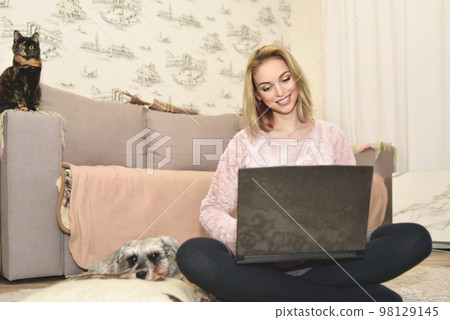Lady smiles during a video call on a laptop, little cat sitting on the couch and cute dog is sleeping on a pillow nearby. 98129145