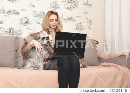 A charming lady is sitting at home with a laptop on the couch and hugging a cute gray dog. 98129146