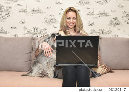 Woman covered with a warm blanket sits on the sofa at home with her dog and smiles happily while chatting by video call on a laptop. 98129147
