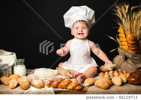 Happy baby boy sits in cook costume among baking bread rolls on the table on black background 98129713