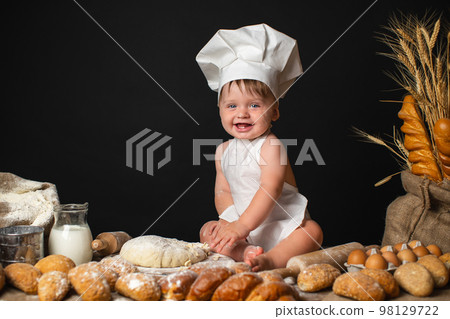 Happy little boy sits in cook costume among baking bread rolls on the table and kneading the dough Happy little boy sits in cook costume among baking bread rolls on the table and kneading the dough 98129722