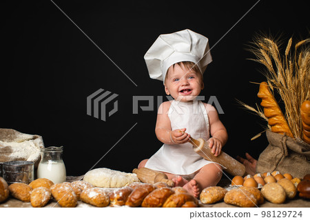Laughing little boy sits in cook costume among baking bread rolls on the table on black background 98129724