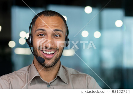 Close up hispanic male worker with video headset smiling and looking at camera, tech support worker working inside office. Close up hispanic male worker with video headset smiling and looking at camera, tech support worker working inside office. 98132734