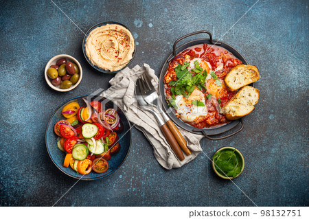 Middle Eastern breakfast or brunch with Shakshouka in pan, toasts, vegetables salad, hummus, olives 98132751