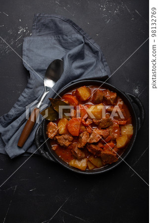 Beef meat stew with potatoes, carrot and delicious gravy in black casserole pot with bay leaves with spoon on black dark rustic concrete background from above . 98132769